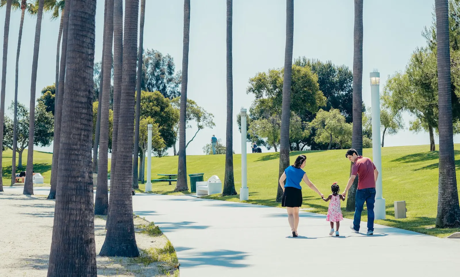Parents walking hand in hand with their young daughter along a palm-lined path