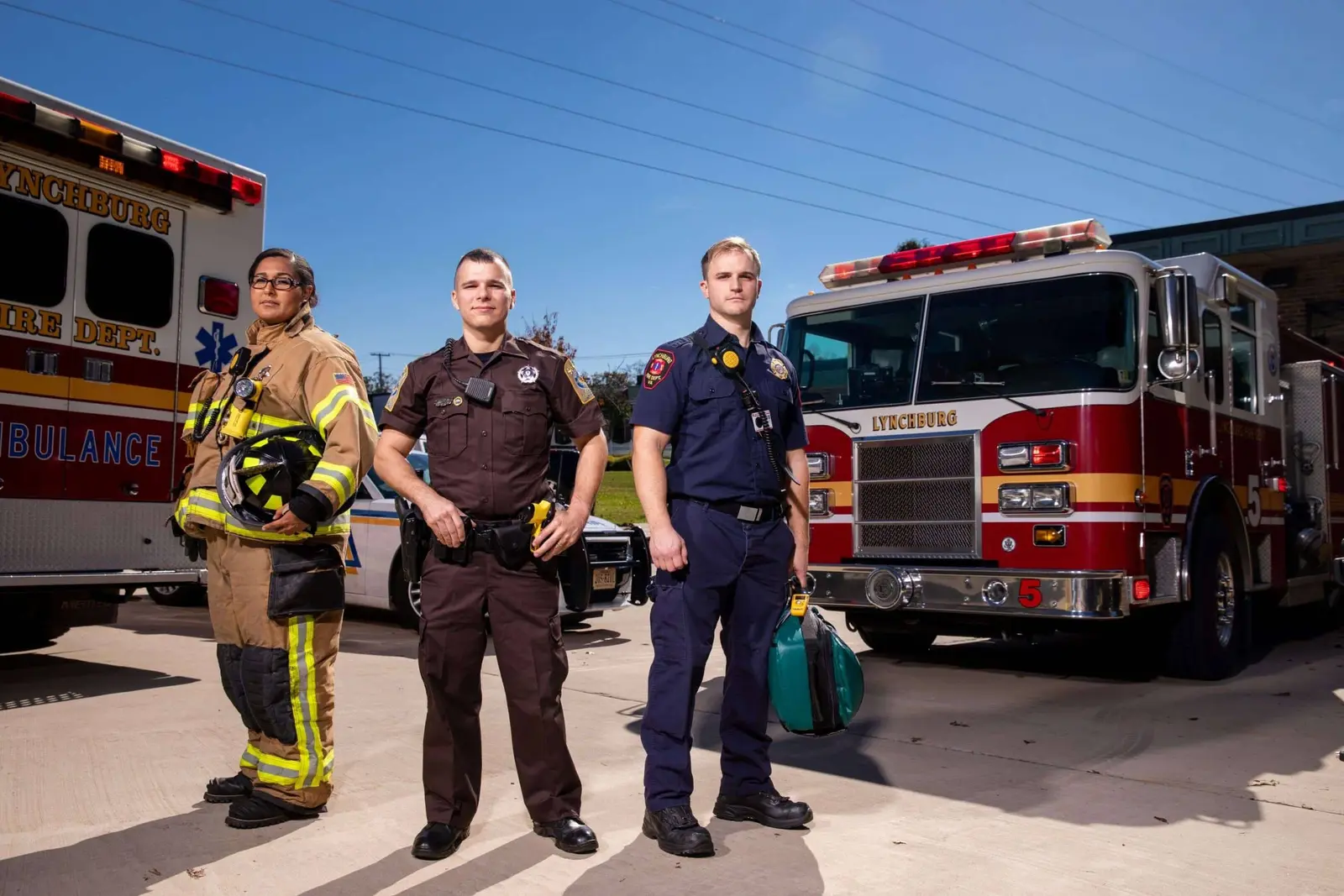 A firefighter, police officer, and EMT standing in front of their vehicles