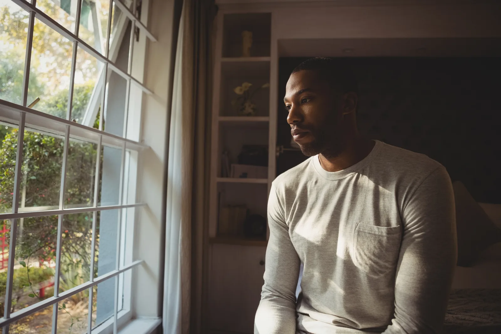 A man seated beside a bedroom window looks outside.