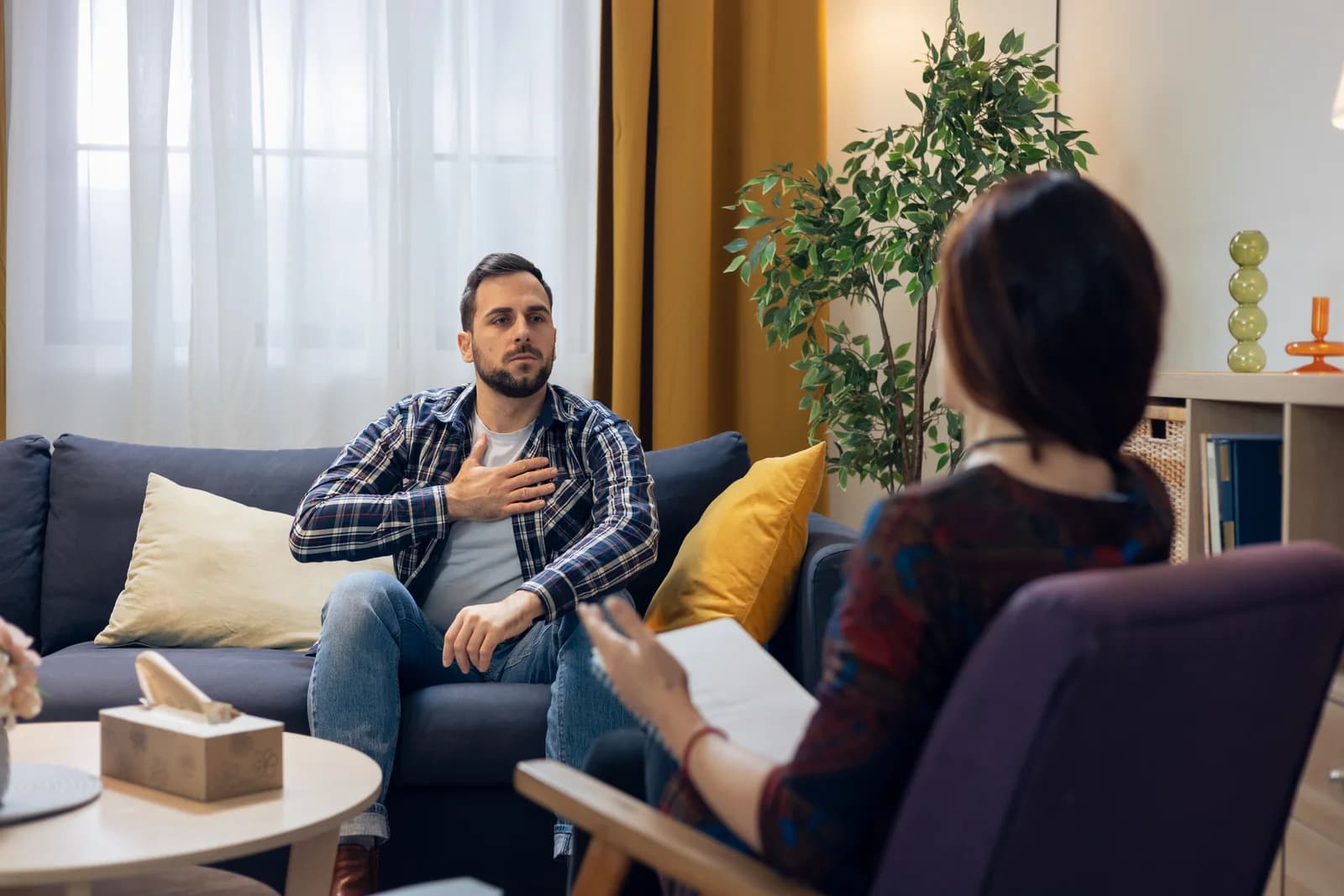 A man seated on a sofa talks with a therapist who is holding a notebook.