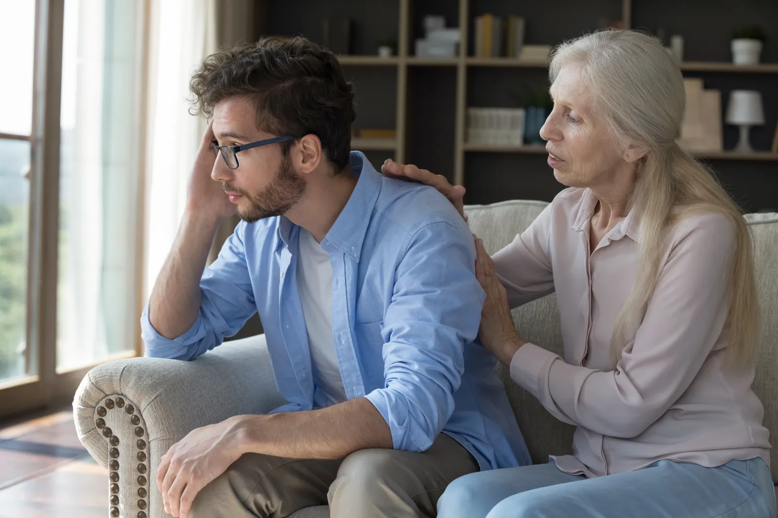 An older woman places a hand on the shoulder of her distressed adult son as they sit on a sofa.