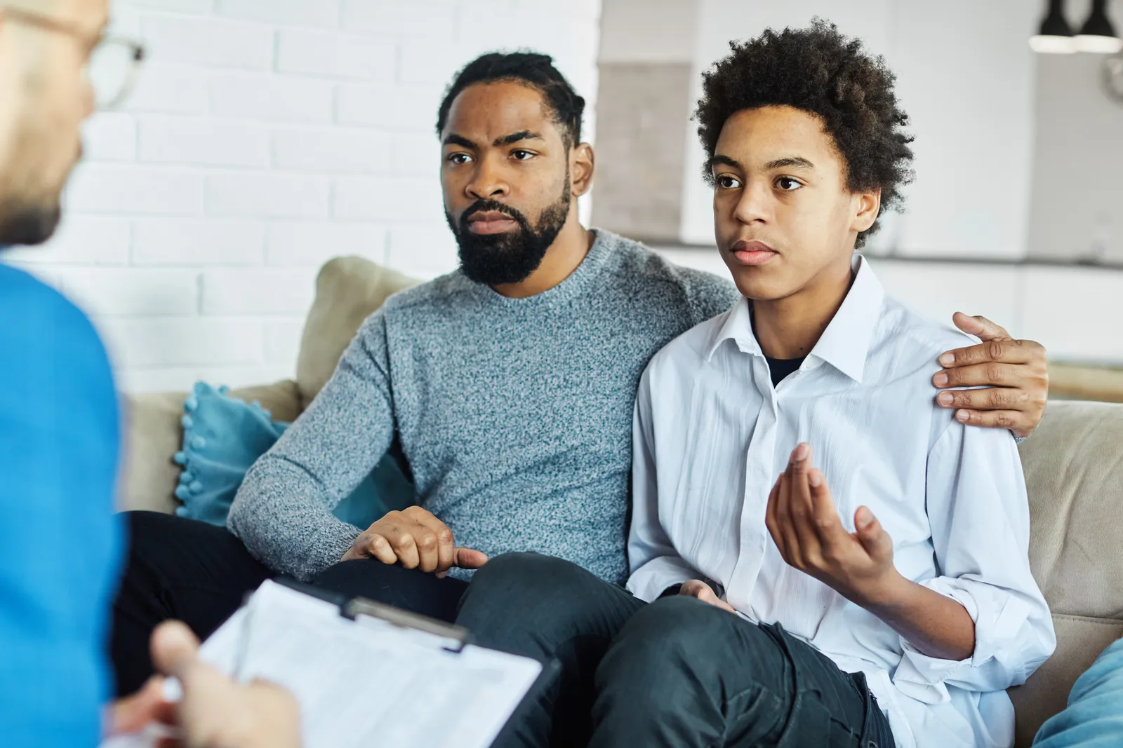 A father with his arm around his teenage son as they talk with a counselor holding a clipboard.
