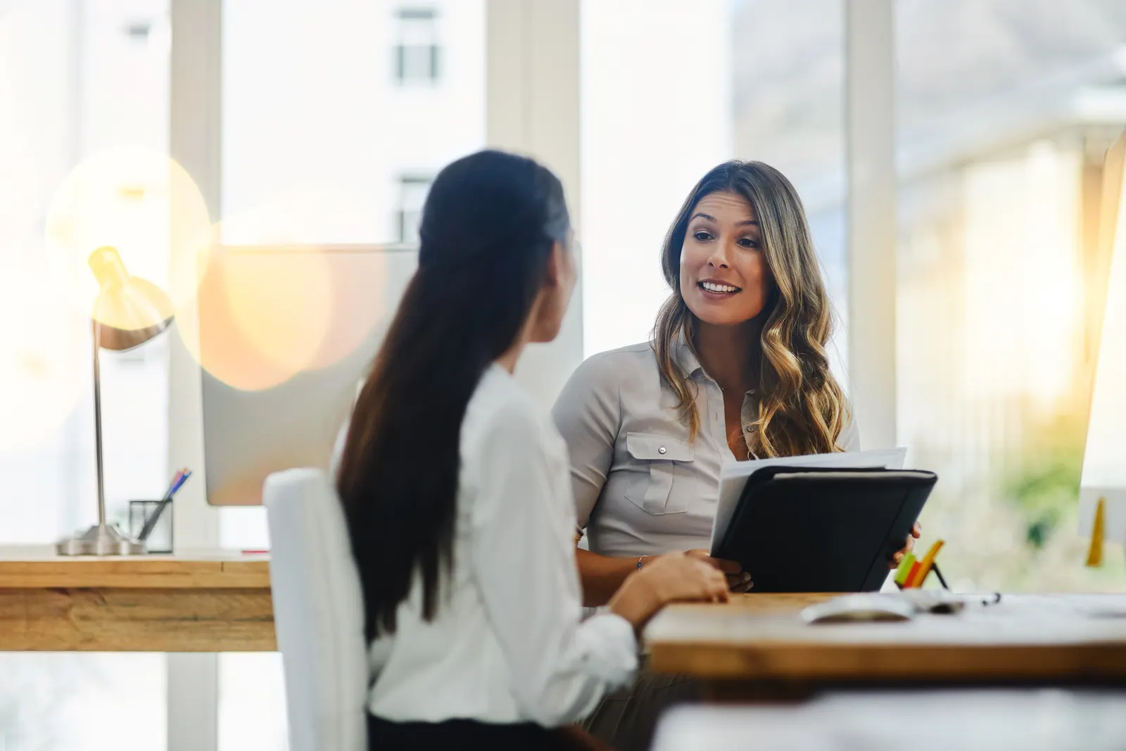 Two women talk across a desk in a bright office, one holding a tablet.