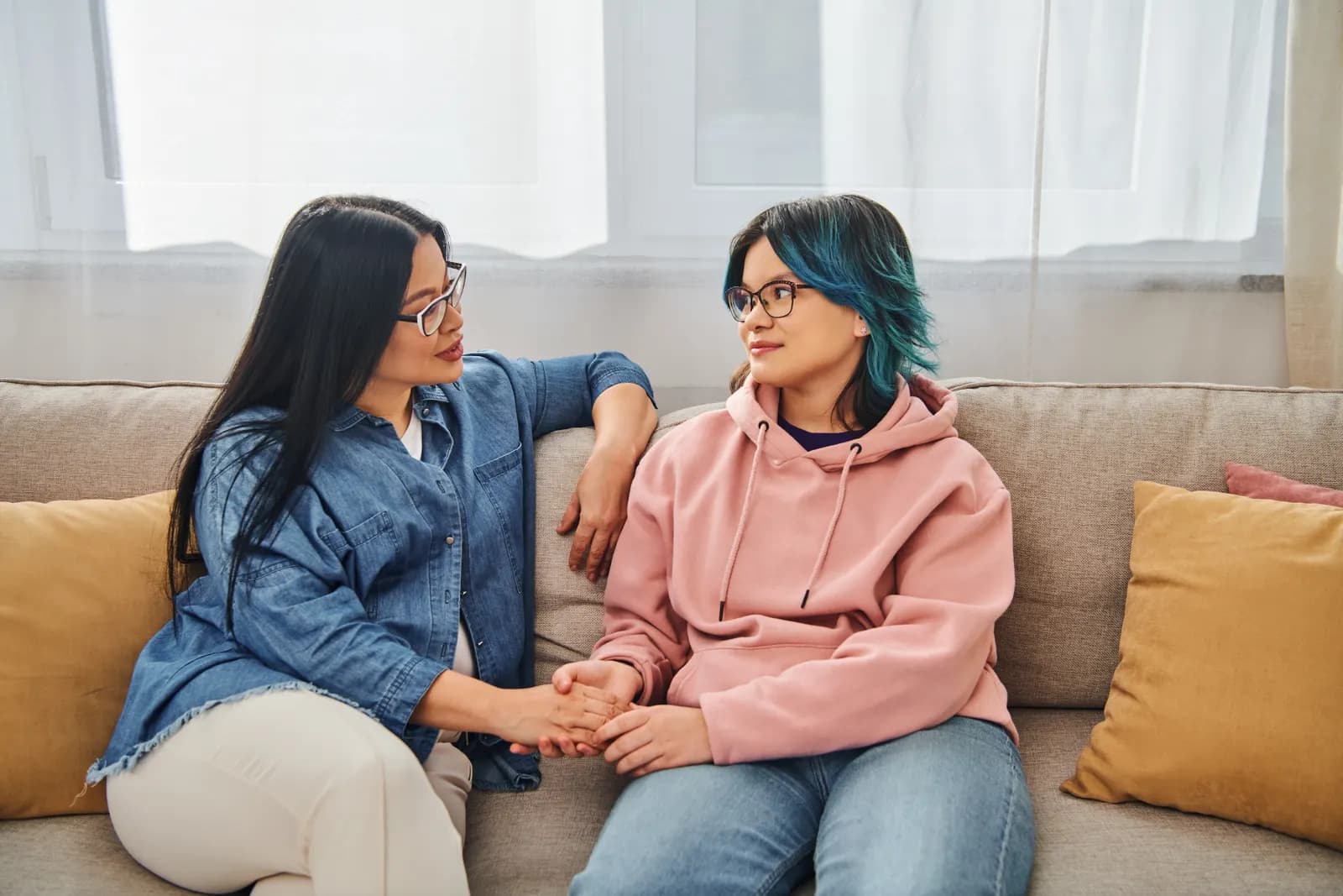 A mother holds her teenage daughter's hands as they talk on a living room couch.